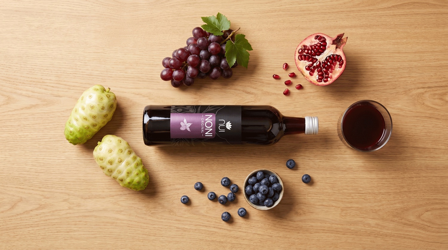 NUI NONI Original: Flasche und Glas zwischen Trauben, Granatapfel, Heidelbeeren und Noni-Früchten